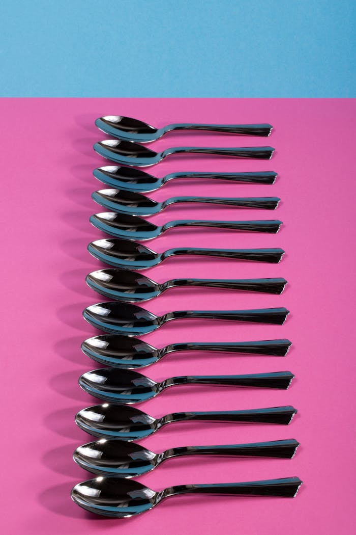 Twelve shiny silver spoons neatly arranged on vibrant pink and blue background.